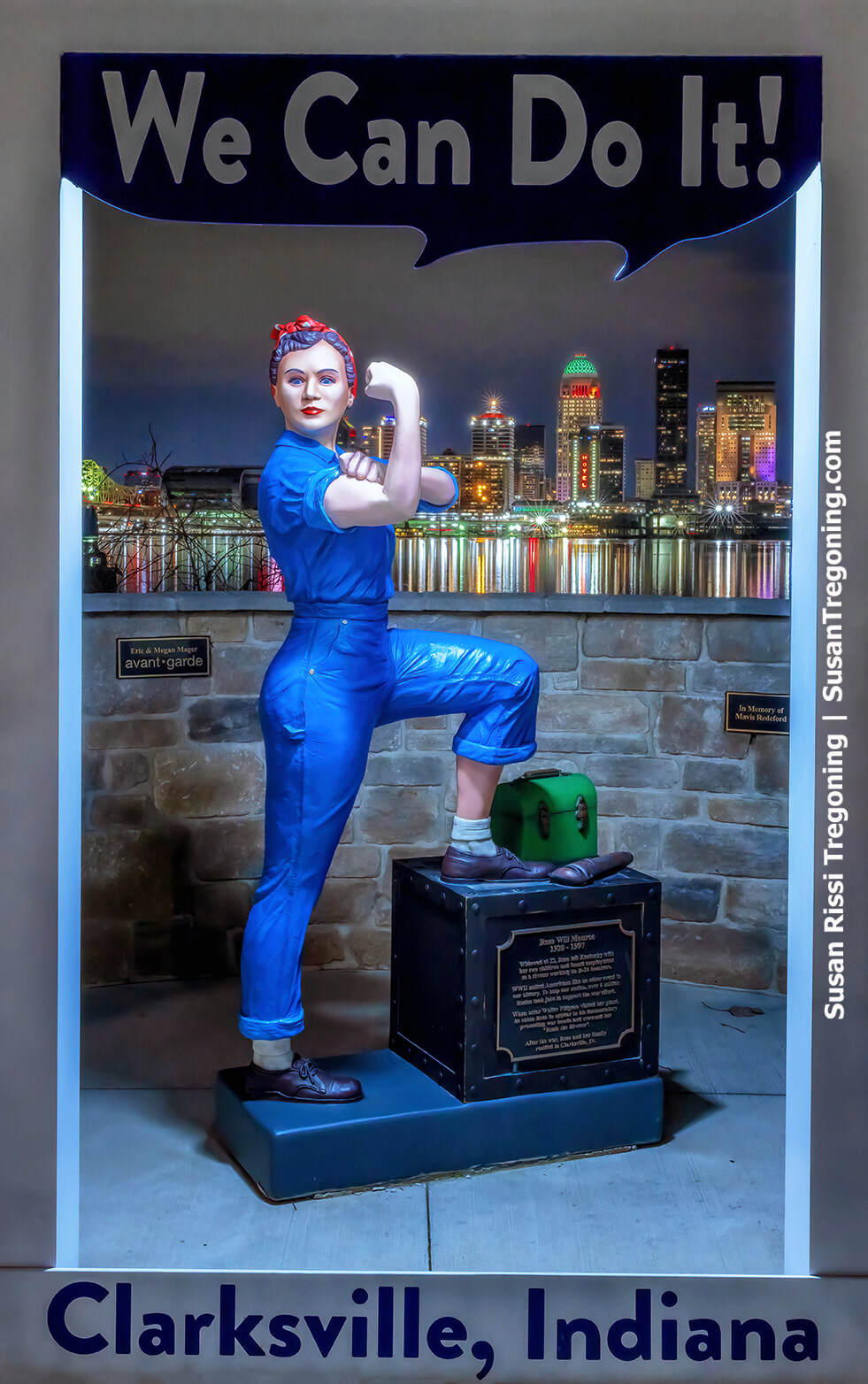 A life‑size painted statue of Rosie the Riveter stands outdoors in a confident pose, with one arm raised in a flexing gesture. The figure wears a blue work shirt, rolled‑up sleeves, and a red‑and‑white polka‑dot headscarf, representing the iconic World War II symbol in Clarksville, Indiana.