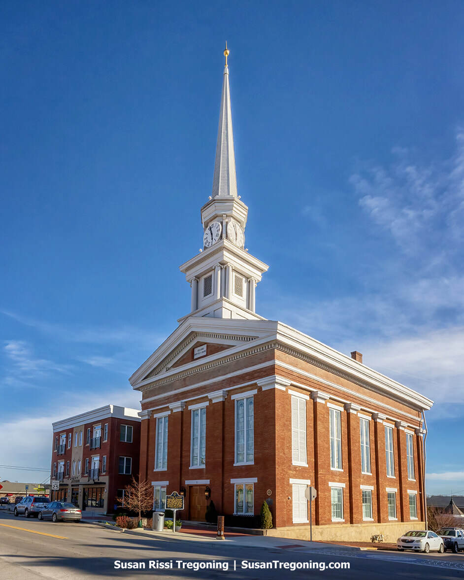 A tall brick church tower with a large clock face rises against the sky, its pointed spire topped with a cross. The building’s windows and decorative brickwork highlight its historic architecture, and bare tree branches frame the scene.