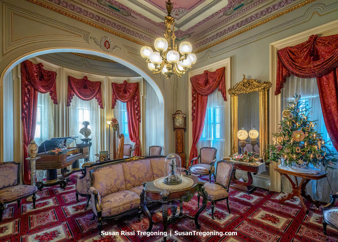 An ornate Victorian parlor features patterned wallpaper, carved wood trim, and an elaborate fireplace with a large mirror above it. A crystal chandelier hangs from the ceiling, and antique furniture--including upholstered chairs and a small table--fills the richly decorated room.