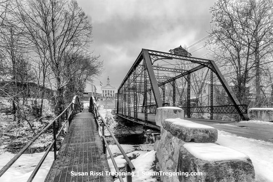  A historic iron truss bridge spans a snowy creek, its metalwork marked with a “Phoenix Iron Co. Phoenixville PA” plaque. A narrow pedestrian walkway runs alongside the bridge, and a domed courthouse with flags rises in the background among bare winter trees.