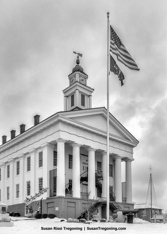 A historic courthouse with tall white columns and a central cupola stands in a snowy winter scene. An American flag flies on a pole in front of the building, and a metal staircase is attached to the side. A Christmas tree‑shaped light display is visible in the background beneath an overcast sky.