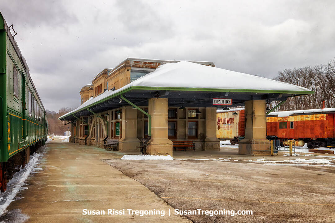 A historic train station with stone pillars and a snow‑covered roof displays a sign reading “French Lick.” Benches sit beneath the covered platform, and several railcars--including a green passenger car and a white car marked “Pluto Water”--stand on the tracks beside the station on an overcast winter day.
