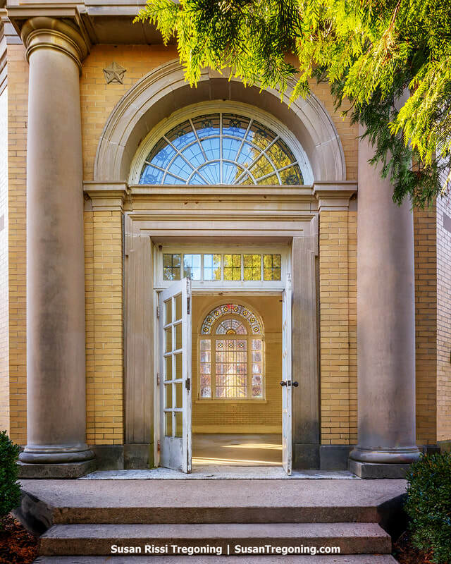 The entrance of a historic brick building with large stone columns frames an open doorway, revealing an interior arched stained‑glass window. A six‑pointed star is set into the brickwork above the door, and tree branches partially frame the scene from the upper right.