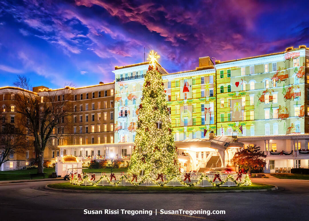 The historic French Lick Springs Hotel in French Lick, Indiana, is illuminated with colorful Christmas-themed light projections, including Santa and ornaments. In front of it stands a tall Christmas tree decorated with white lights and topped with a glowing star, surrounded by a lit circular fence and festive bows. The sky is deep purple and blue, adding to the dramatic holiday atmosphere.