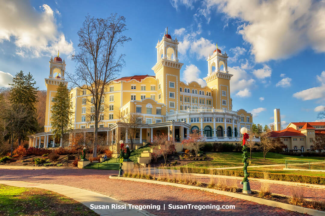  A large historic hotel painted yellow with white trim stands under a partly cloudy sky. The multi‑story building features several towers topped with red domes and a wide columned porch across the front. Manicured landscaping, including trees and shrubs, surrounds a brick-paved drive leading to the entrance. Sunlight highlights the architectural details.