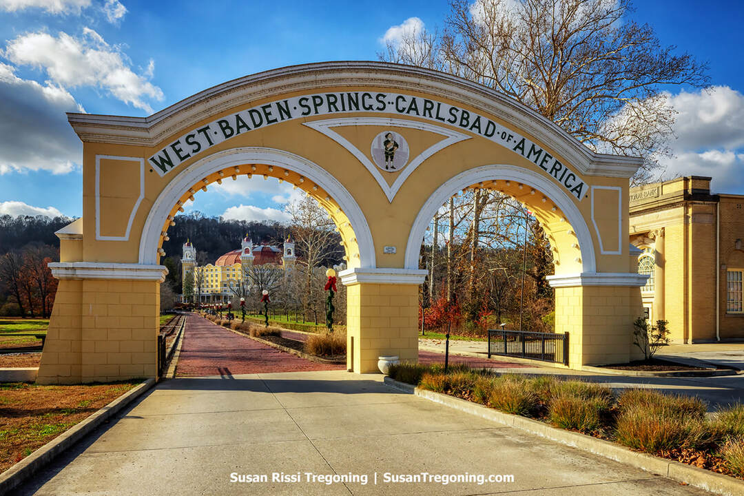 An ornate yellow-and-white arched gateway labeled “West Baden Springs – Carlsbad of America” stands over a red brick drive. The central arch is the tallest, topped with a decorative emblem. Beyond the gateway, a domed historic West Baden Springs Hotel rises among bare trees under a bright sky with scattered clouds.