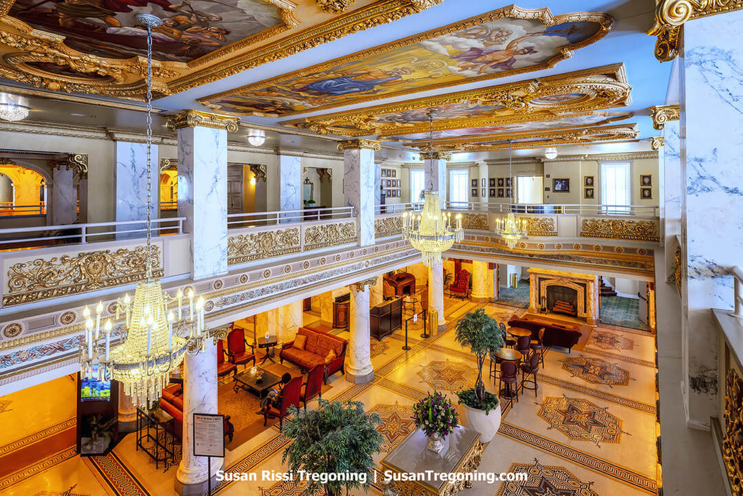 The Grand Lobby of the historic French Lick Springs Hotel and Spa, with marble columns, ornate gold detailing, and large crystal chandeliers, features red upholstered chairs, wooden tables, potted plants, and floral arrangements. A decorated ceiling with painted murals and gold-leafed frames, as seen from the second-floor balcony, overlooks the space.