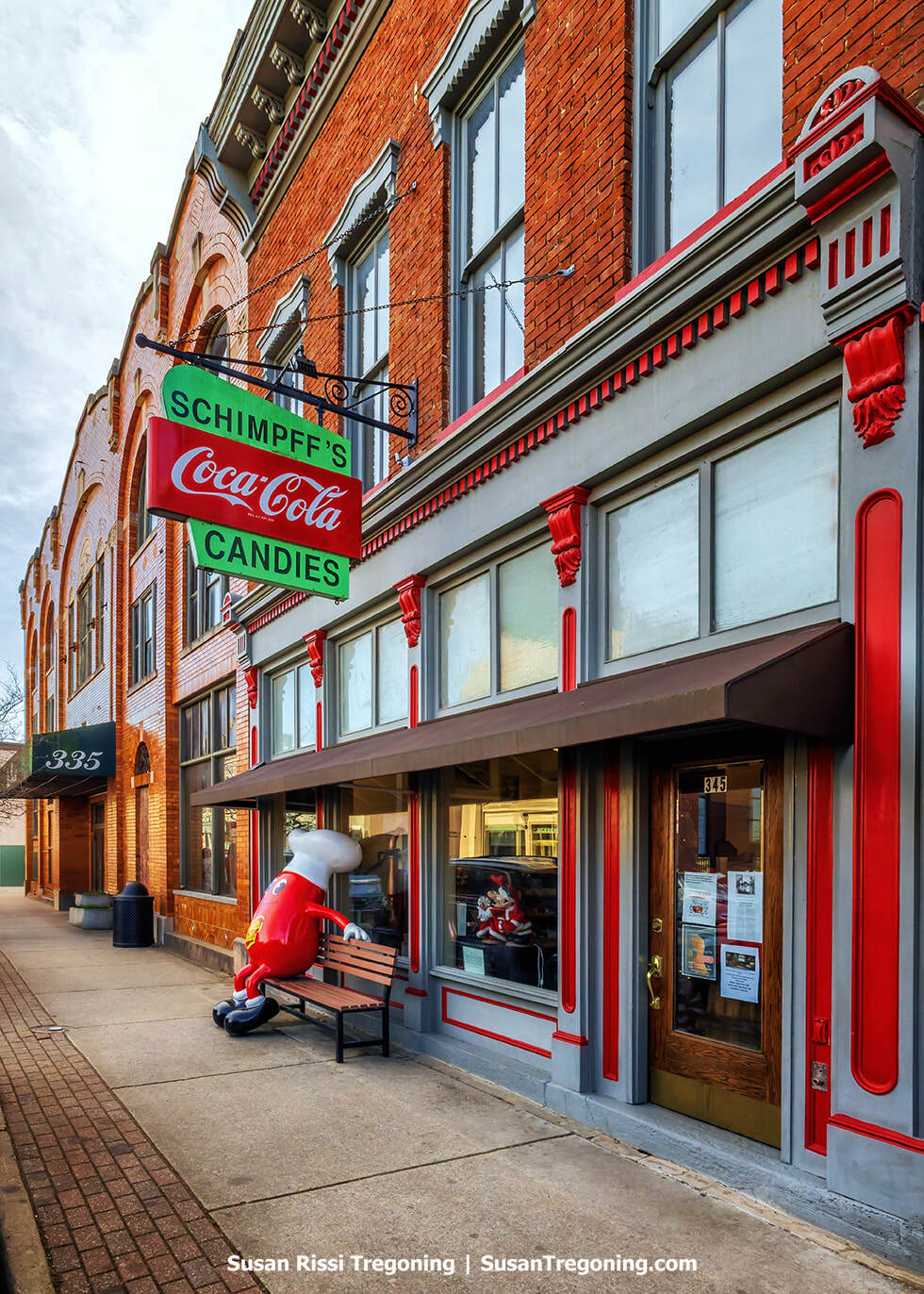 The historic storefront of Schimpff’s Confectionery features a brown awning above large display windows filled with vintage candy jars and old‑fashioned sweets. The brick building sits along the sidewalk in downtown Jeffersonville, creating a nostalgic small‑town streetscape.