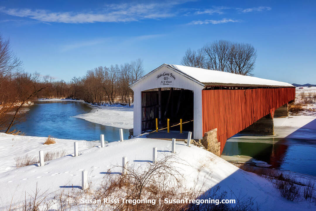 A serene winter view of the historic Medora Covered Bridge in Indiana, captured with snow-covered landscapes and crisp blue skies showcasing the charm of this 1875 landmark.