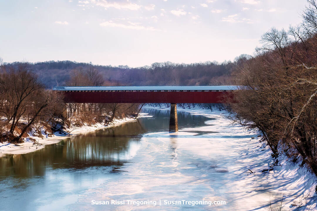 The Williams Covered Bridge in Lawrence County, Indiana, stretches across the partly iced East Fork of the White River on a frigid, snow‑covered day. The long red bridge stands above a central stone pier, with bare winter trees lining both banks under soft, partly cloudy light.