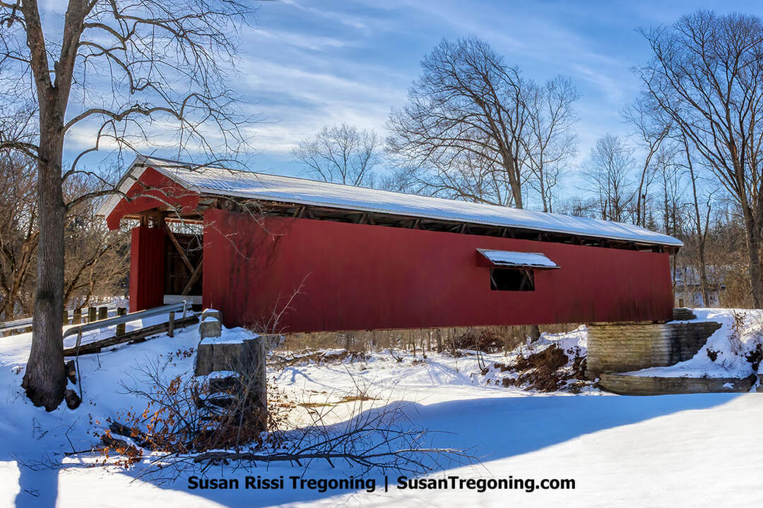 A historic red covered bridge known as the Otter Creek Covered Bridge stands broadside on a snowy winter day near Holton, Indiana. The wooden Howe‑truss span crosses Otter Creek and is surrounded by leafless trees and fresh snow.