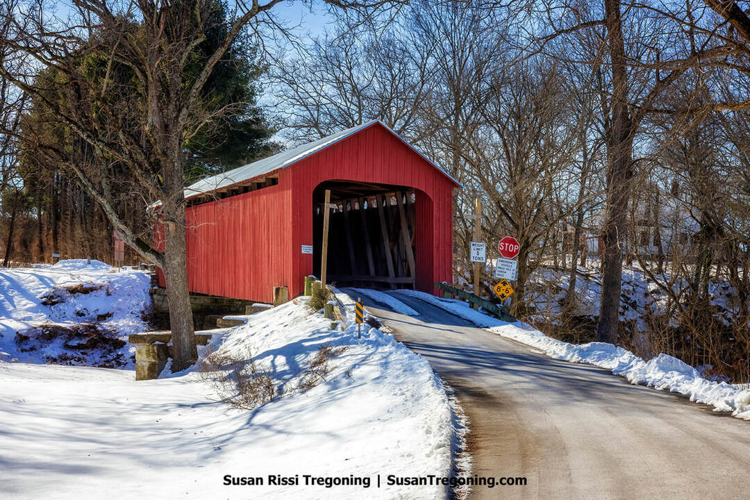 A red covered bridge stands at the end of a snow‑covered road, surrounded by leafless winter trees. Several road signs--STOP, a 3‑ton weight limit, and a 6′‑6″ clearance sign--are posted near the entrance. The scene is quiet and wintry