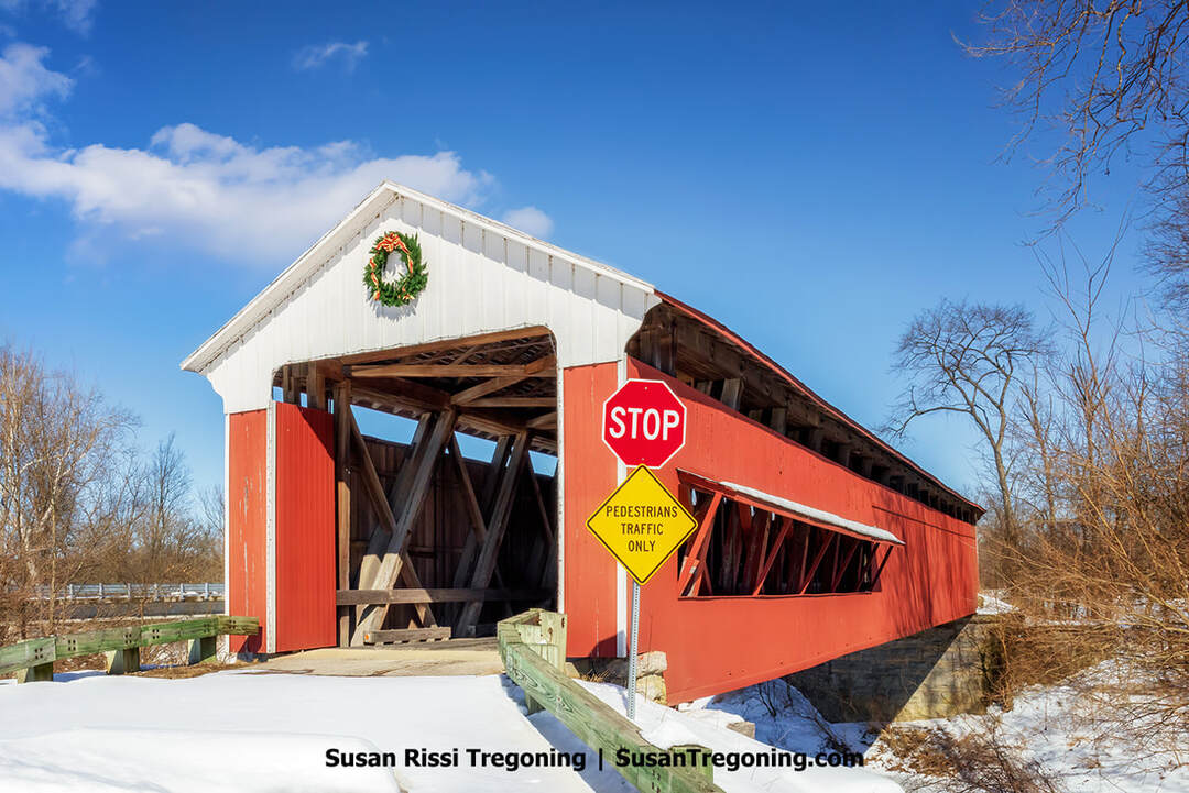 A red-and-white historic Scipio Covered Bridge in Jennings County, Indiana, decorated with a holiday wreath, stands in a snowy winter landscape, surrounded by bare trees under a bright blue sky.
