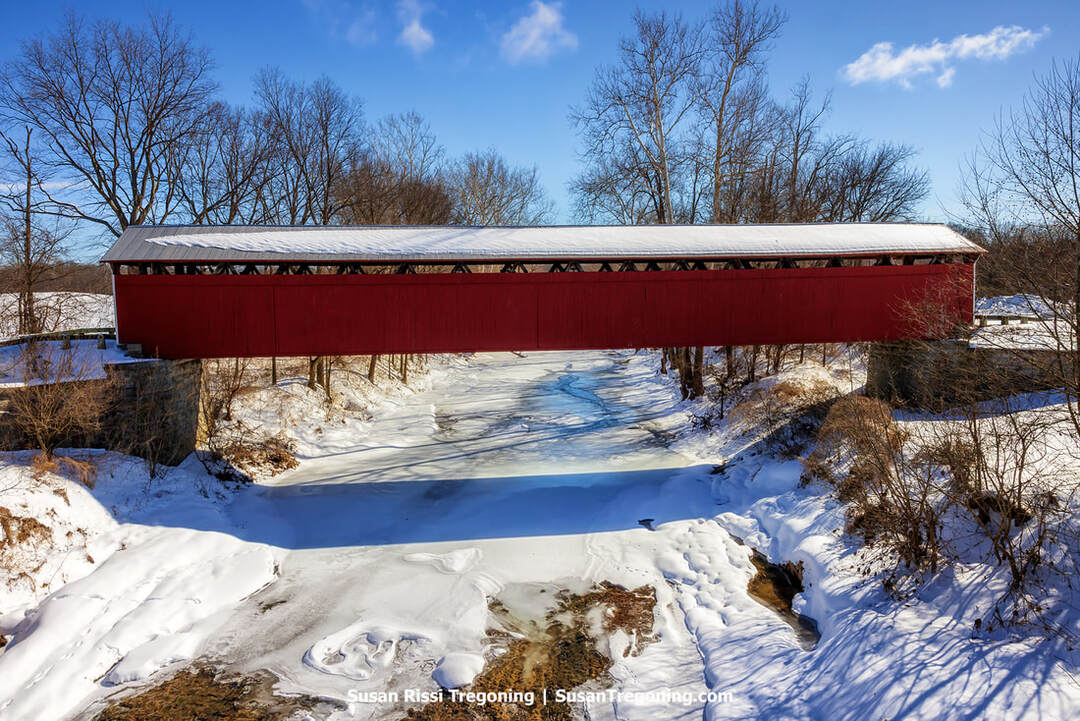 The historic red Scipio Covered Bridge in Jennings County, Indiana, shown broadside across the frozen Sand Creek, set in a bright, snow-covered winter landscape with leafless trees and clear sunlight.