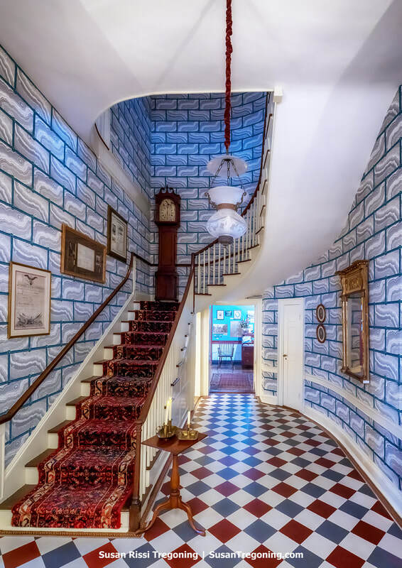 The entry hall of Grouseland features a curved staircase with a red patterned carpet runner rising to the right. A tall grandfather clock stands on the staircase landing before the first turn. The walls are covered in blue‑and‑white faux‑brick wallpaper, and the floor is tiled in a red, white, and blue‑gray checkerboard pattern. Framed artwork and documents hang on the walls, and a small wooden table with decorative items sits near the staircase. A hanging light fixture illuminates the hallway, which opens into an adjoining room.