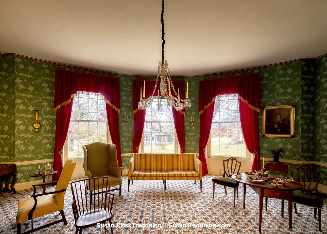 The formal parlor at Grouseland features green patterned wallpaper, red draperies with gold trim, and a crystal chandelier hanging above period furniture. A striped settee, upholstered chairs, and a wooden table set with fine china are arranged on a patterned carpet. A portrait in a gold frame hangs on the wall, and a wall clock is visible to the left. Large windows bring natural light into the historic room.