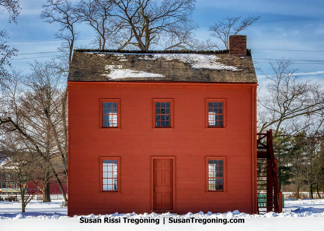 The Indiana Territorial Capitol in Vincennes, known as the “Red House,” is a two‑story red‑painted wooden building standing in a snowy landscape. The structure has a symmetrical front with five windows and a central door, and an exterior staircase on the right side leads to the second floor. Snow covers the ground and parts of the roof, and leafless trees surround the historic building beneath a bright winter sky.