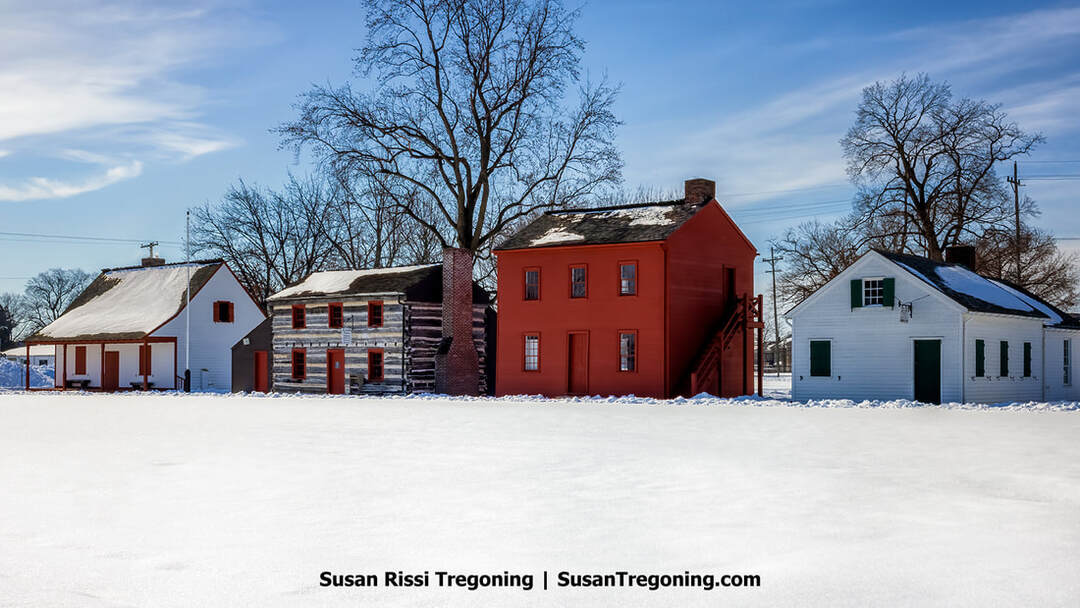 A row of historic buildings at the Vincennes State Historic Sites stands in a snowy landscape under a bright blue sky. The structures include the red-painted Old Indiana Territorial Capitol, along with white frame buildings and a log cabin. Leafless trees rise behind the buildings, and snow covers the ground and rooftops, creating a quiet winter scene.