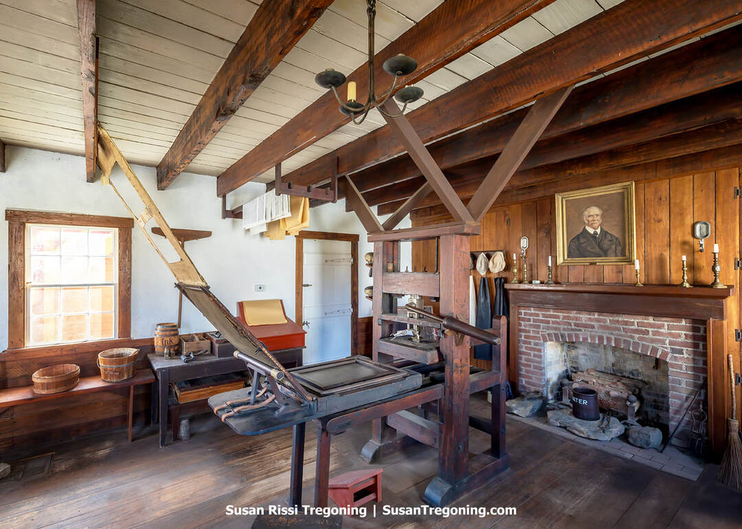 Inside the Elihu Stout Print Shop at the Vincennes State Historic Sites, a large wooden Adam Ramage printing press stands at the center of the room. Printing tools, paper, and type cases surround the press. A brick fireplace with candlesticks and a framed portrait sits to the right, with garments and hats hanging nearby. Wooden beams, rustic furnishings, and soft light create the atmosphere of an early American print shop.