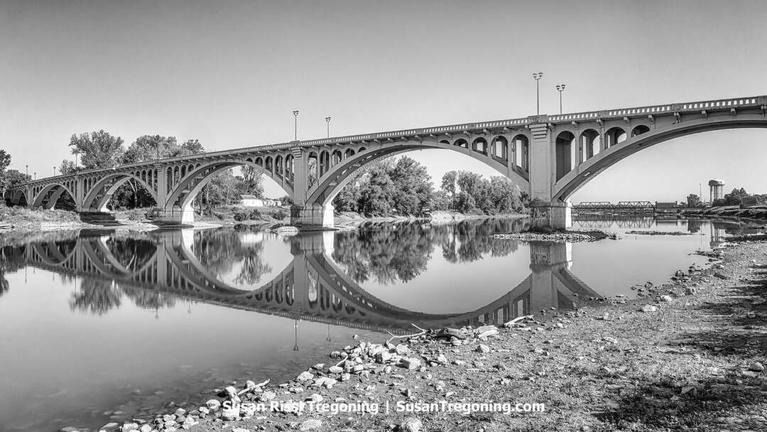 A black‑and‑white photograph of a multi‑arched concrete bridge spanning a calm river, its arches and railings reflected clearly in the still water. Streetlights line the top of the bridge, and trees frame both banks. A rocky shoreline appears in the foreground, with a smaller bridge and a distant water tower visible in the background. 