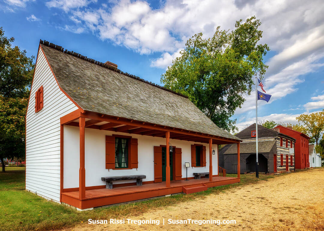 Jefferson Academy, a white wooden building with deep orange trim and a steeply pitched roof, stands on a grassy lawn at the Vincennes State Historic Sites. The structure has shuttered windows, a front porch with benches, and a green door. Behind it are additional historic buildings, including a log cabin and a red-painted structure. A flagpole with the American flag rises nearby, and trees and a partly cloudy sky frame the scene.