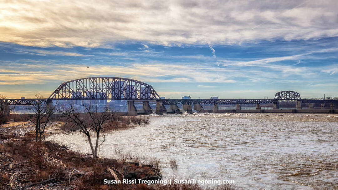 A long steel railroad bridge spans the Ohio River, its dark truss structure stretching toward Louisville. High water covers the normally exposed fossil beds, with only a few rocks visible near the shoreline in the foreground.