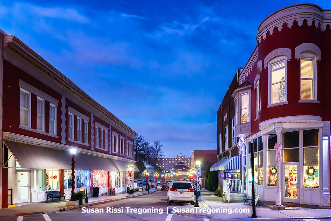 A small‑town main street at dusk features historic red‑brick buildings decorated with holiday wreaths, ribbons, and lights. Storefront windows glow warmly, a few parked cars line the street, and illuminated streetlamps brighten the scene beneath a deep blue evening sky.