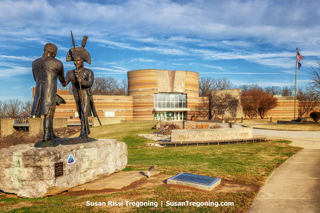 A life‑size statue of Meriwether Lewis and William Clark stands in the foreground, depicting the two explorers shaking hands at their first meeting. Both figures wear period frontier clothing. Behind them, the modern Falls of the Ohio Interpretive Center rises with its angular rooflines and large windows, marking the historic site where they joined forces at George Rogers Clark’s cabin before beginning their expedition.