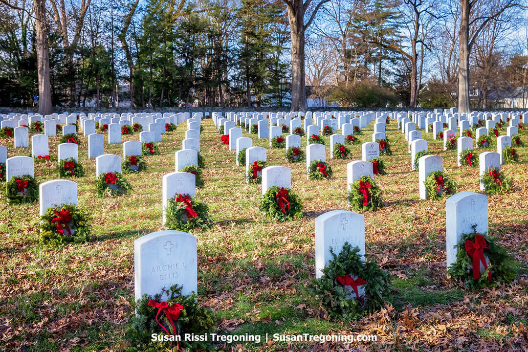Rows of white military headstones stand in neat lines across the cemetery, with green Christmas wreaths placed at the base of each marker. Leafless trees and an American flag are visible in the background.