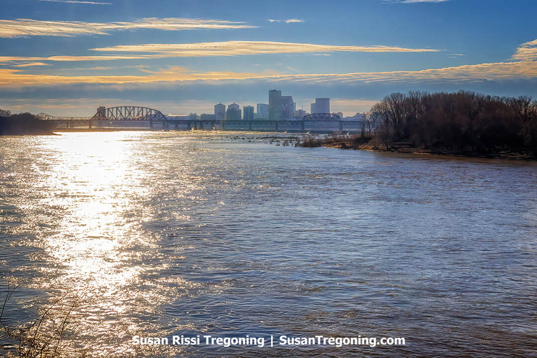 A view from the George Rogers Clark homestead site overlooks the Ohio River, with the stone channel wall running along the foreground and a tree‑covered island positioned mid‑river. The water stretches beyond the island toward the far shoreline, creating a calm, expansive river scene.