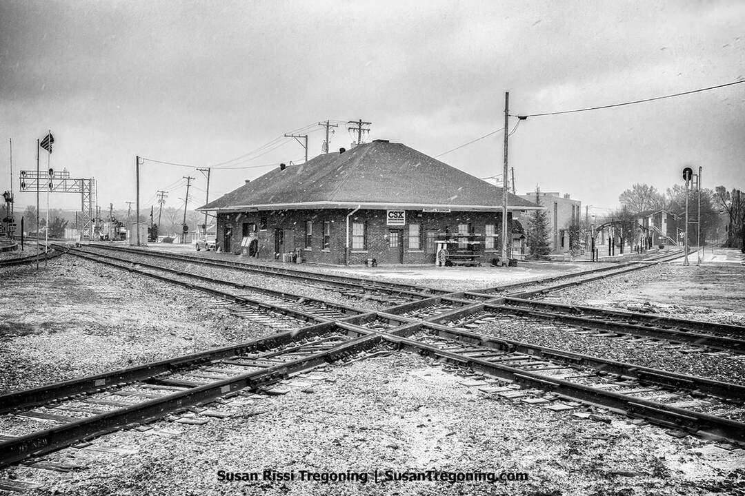 Black‑and‑white photo of snow falling over the diamond railroad crossing in North Vernon, Indiana, with multiple intersecting tracks leading toward a brick building labeled ‘CSX Transportation.’ Utility poles, signal lights, and small town buildings appear in the background.