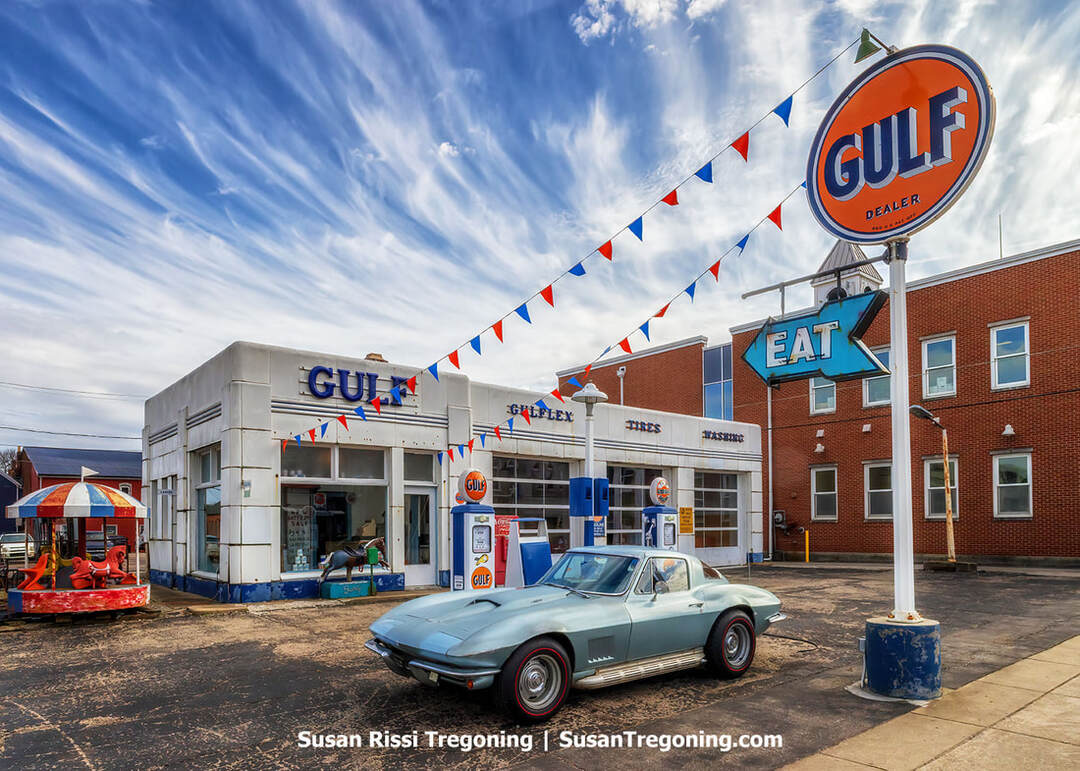 A vintage 1940s Gulf gas station in Aurora, Indiana, with white walls and blue trim, stands with a classic 1963 Corvette Stingray parked at the pump. Red and blue pennant flags hang across the lot, with a brick building and an “EAT” sign to the right and a small carousel to the left under a sky of wispy clouds.