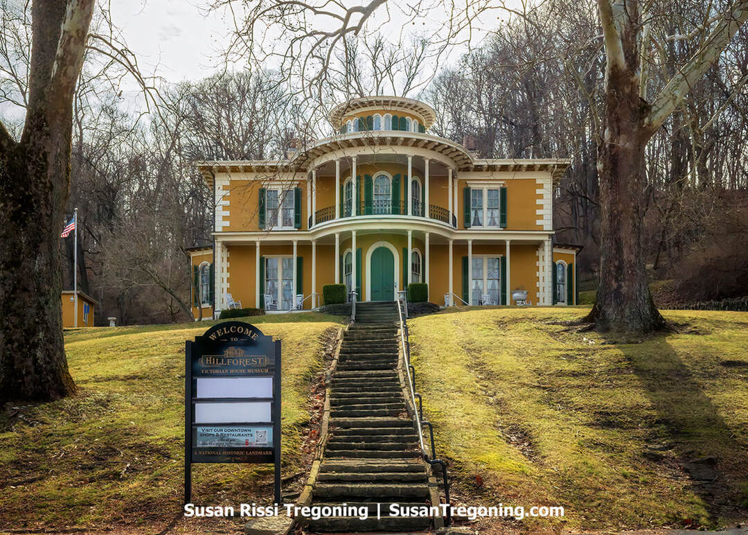 The two-story yellow Italianate Hillforest Victorian House Mansion, with a central rotunda, sits high on a grassy hill, reached by a stone staircase. A black “Welcome” sign stands at the base of the steps, and tall leafless trees surround this Steamboat Gothic Historic Landmark in Aurora, Indiana. 