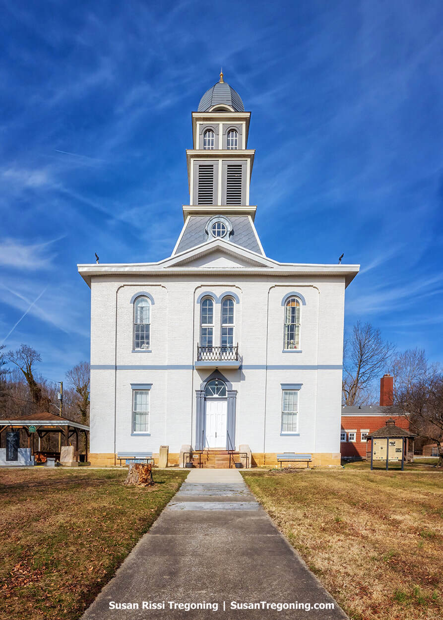 The historic 1876 Martin County Courthouse in Shoals, now home to the Martin County Museum, stands beneath a blue sky with wispy clouds. The white building features a symmetrical façade with arched windows, a central entrance, and a tall multi‑tiered tower with louvered openings and a domed top. A sidewalk leads to the front steps, with benches, a small gazebo‑like structure, and an informational sign on the surrounding lawn.