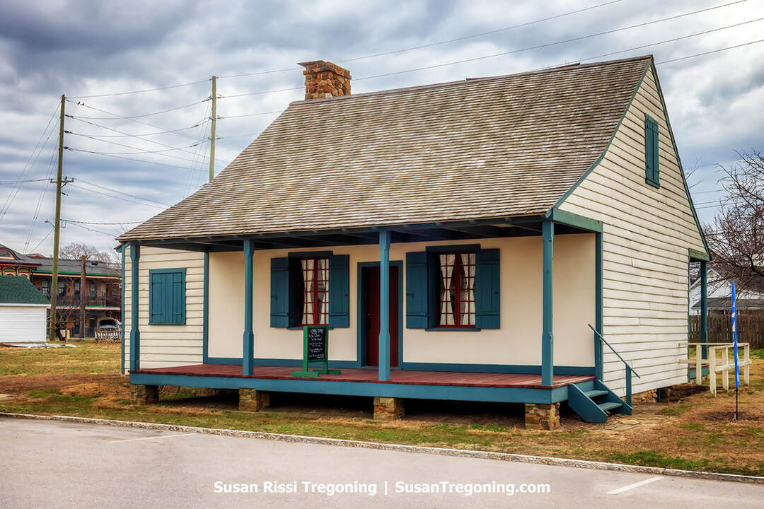 The Old French House, an authentic French Creole residence and the last surviving post‑on‑sill dwelling in Indiana, stands on a raised foundation with a steep roof, blue shutters, and a columned front porch. Nearby buildings and power lines are visible in the background.