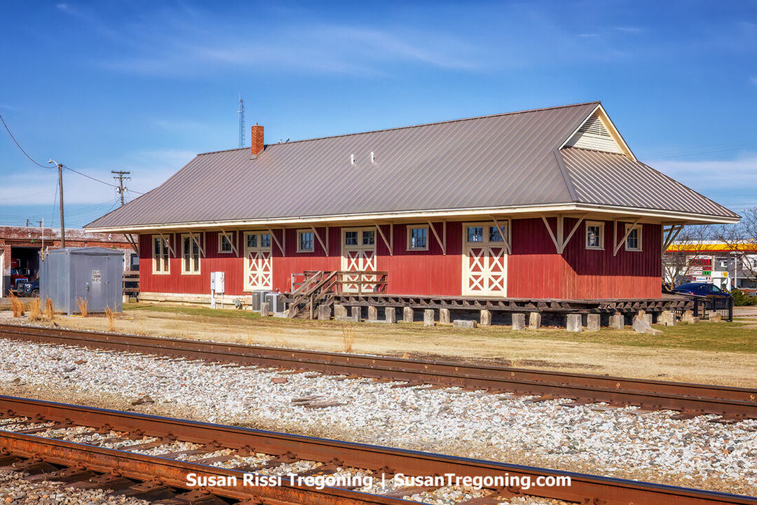 The Southern Indiana Railroad Freighthouse stands beside a set of train tracks on a clear day. The long, low wooden depot has a gabled roof, wide overhangs, and board‑and‑batten siding. A small platform runs along the track side of the building, and leafless trees rise behind it. The scene highlights the structure as the only surviving railroad depot in Seymour.