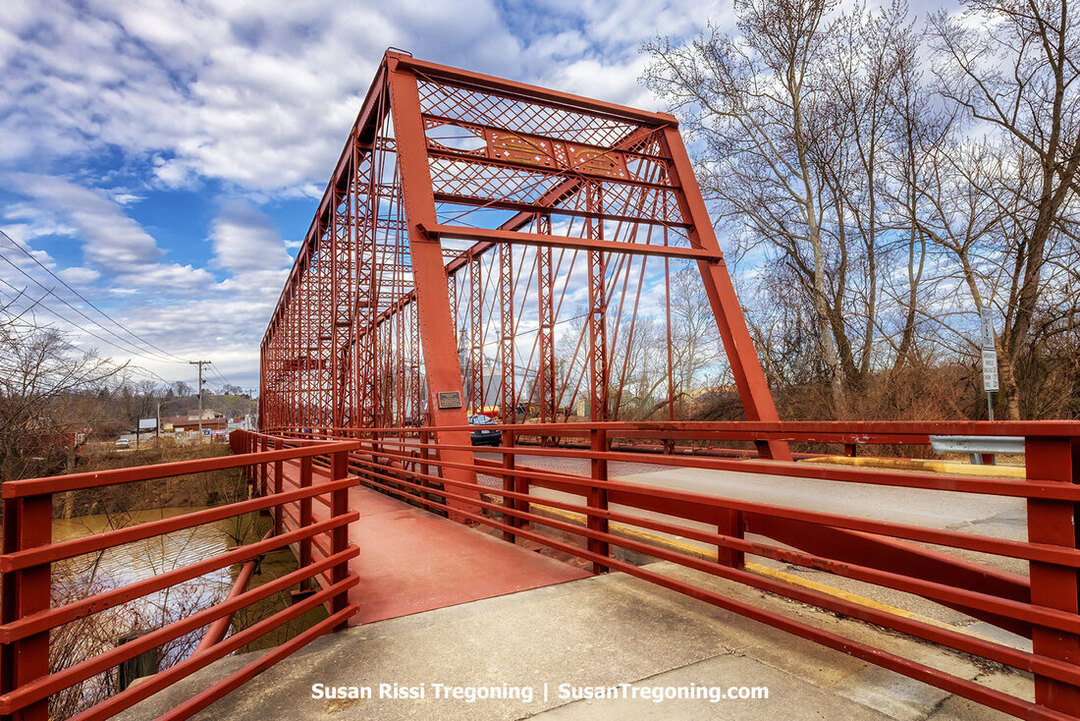 The historic George Street red iron truss bridge spans the Hogan Creek in downtown Aurora, Indiana. A pedestrian walkway on the left and a roadway on the right. Leafless trees line the riverbanks, and a partly cloudy sky appears overhead. A plaque is mounted on one of the vertical beams near the entrance, identifying it as a historic landmark.