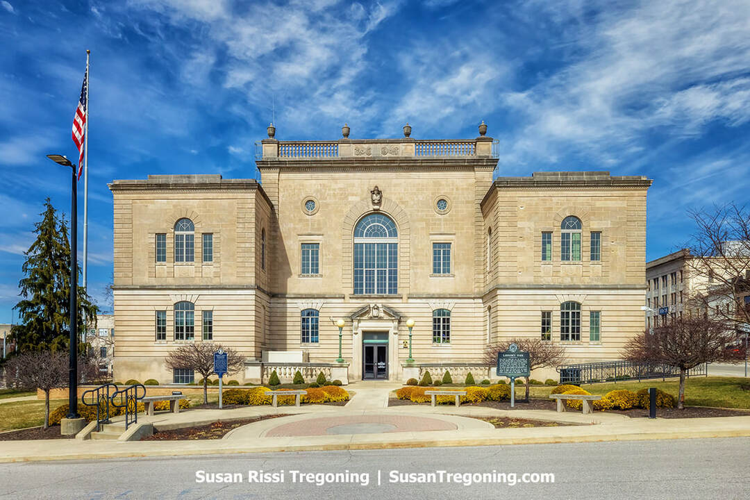 The Lawrence County Courthouse in Bedford, Indiana, appears as a three‑story Neoclassical limestone building with a symmetrical façade, arched windows, and decorative stonework. A central entrance is flanked by lampposts, with landscaped grounds, benches, and a flagpole in front. The courthouse stands beneath a bright sky with scattered clouds.