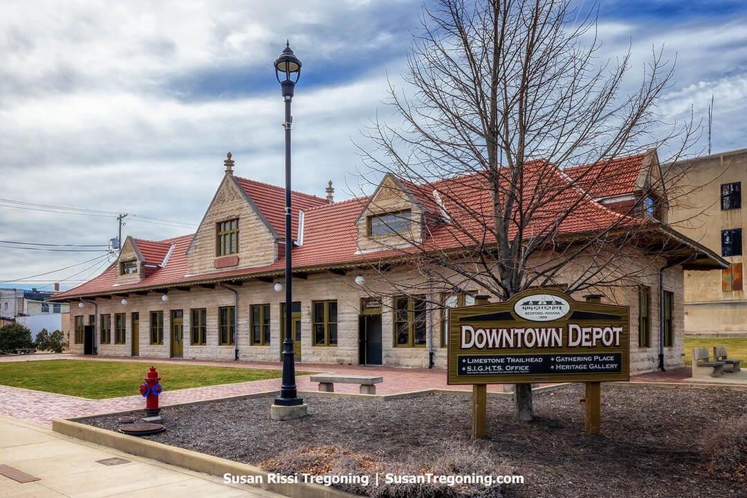 The restored Milwaukee Railroad Depot in Bedford, Indiana, is a single‑story limestone building with a red clay tile roof and gabled sections, shown on a partly cloudy day. A sign in front identifies it as the Downtown Depot and trailhead, with a leafless tree, streetlamp, fire hydrant, and bench in the foreground. Sidewalks and nearby buildings frame the historic structure.