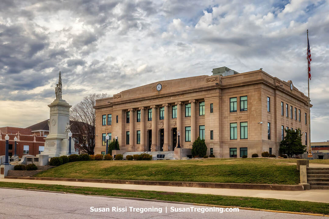 The historic Daviess County Courthouse in Washington, Indiana, stands beneath a partly cloudy sky. The neoclassical building features a symmetrical façade with tall columns, a central clock above the entrance, and large rectangular windows. In front of the courthouse rises a white stone Civil War memorial topped with a statue, and an American flag flies on a tall flagpole to the right.