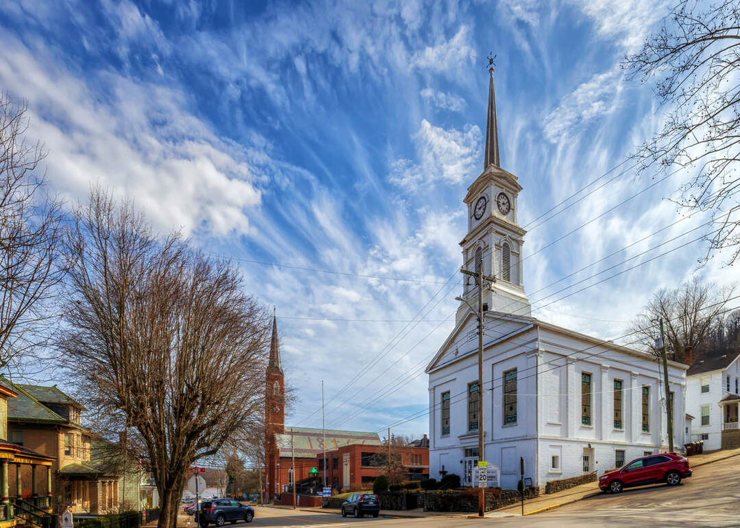 The white First Presbyterian Church, with a tall steeple and clock tower, stands at a sloped street corner in the small town of Aurora, Indiana. Cars are parked along the road, and the red brick St Mary’s Catholic Church with a tall spire rises in the background. Leafless trees and a sky filled with wispy clouds frame the scene.