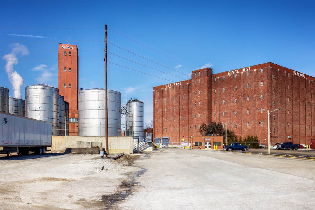 A red brick industrial building with large white “Seagram’s” lettering stands beside several tall metal storage tanks. A red tower with the Ross & Squibb Distillery name rises in the background, and a semi truck is parked along the paved road at the MGP distillery in Lawrenceburg, Indiana.