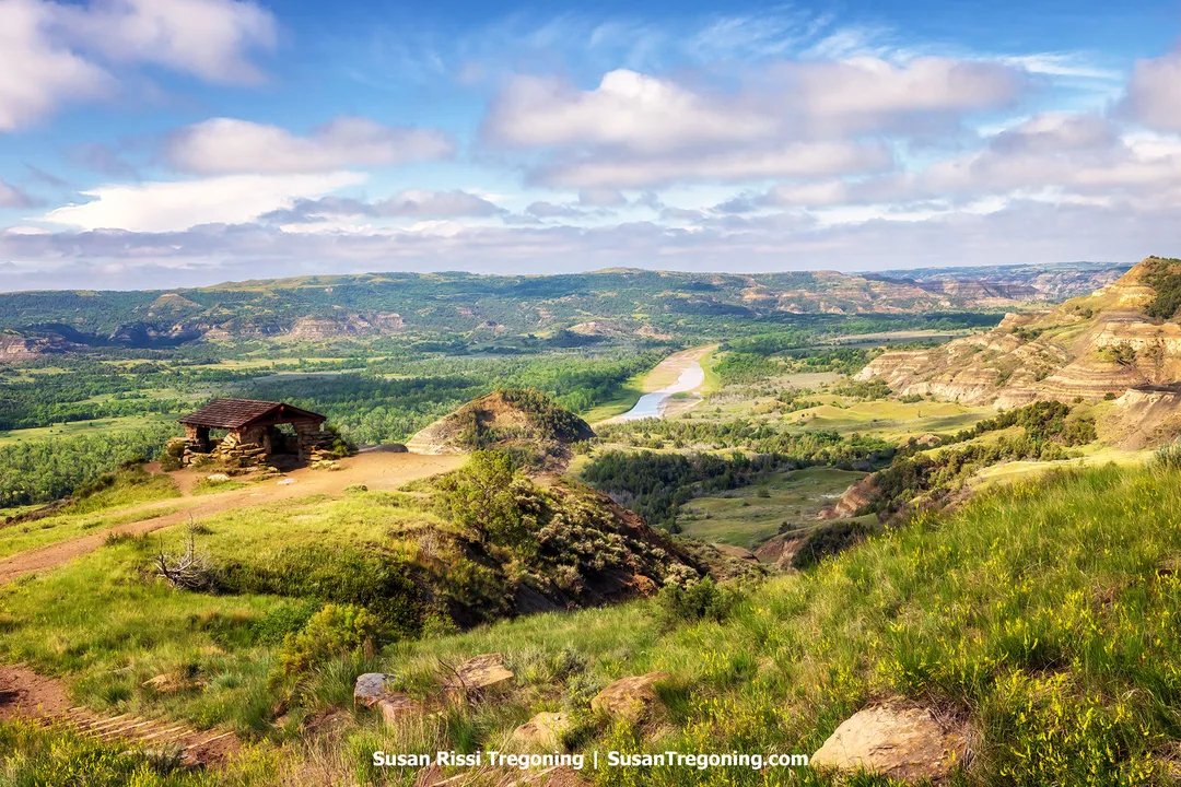 Scenic view of Theodore Roosevelt National Park with a historic CCC-built shelter on a grassy hill overlooking the Little Missouri River and the distant Badlands in early morning light under a partly cloudy sky.