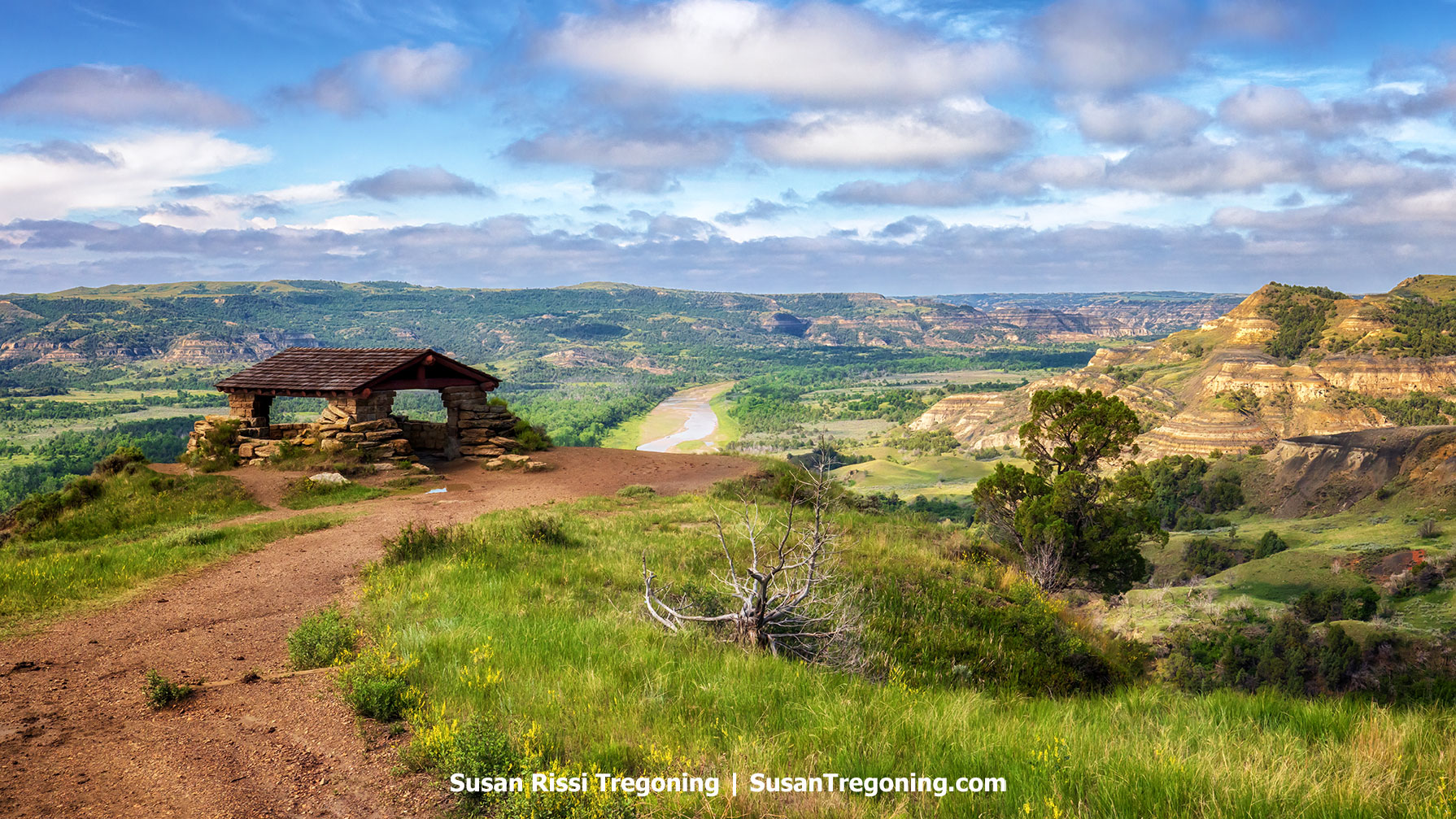 Scenic overlook at Theodore Roosevelt National Park in the North Dakota Badlands, featuring a rustic stone shelter on a grassy bluff with sweeping views of layered hills, winding Little Missouri River, and a vibrant summer sky.