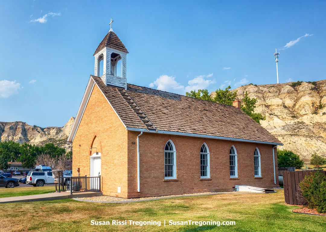 St. Mary’s Catholic Church in Medora, North Dakota, built in 1884 by the town’s namesake Medora von Hoffman, stands beneath the rugged Badlands bluffs. Made from locally crafted brick with a white bell tower and cross, the church reflects frontier faith and aristocratic influence in the early Badlands.