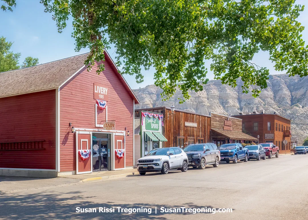 Historic storefronts along 3rd Avenue in Medora, North Dakota, feature the red 1905 Livery Stable adorned with patriotic bunting. Restored wooden buildings line the quiet avenue beneath the rugged backdrop of the Badlands, preserving the town’s frontier charm.