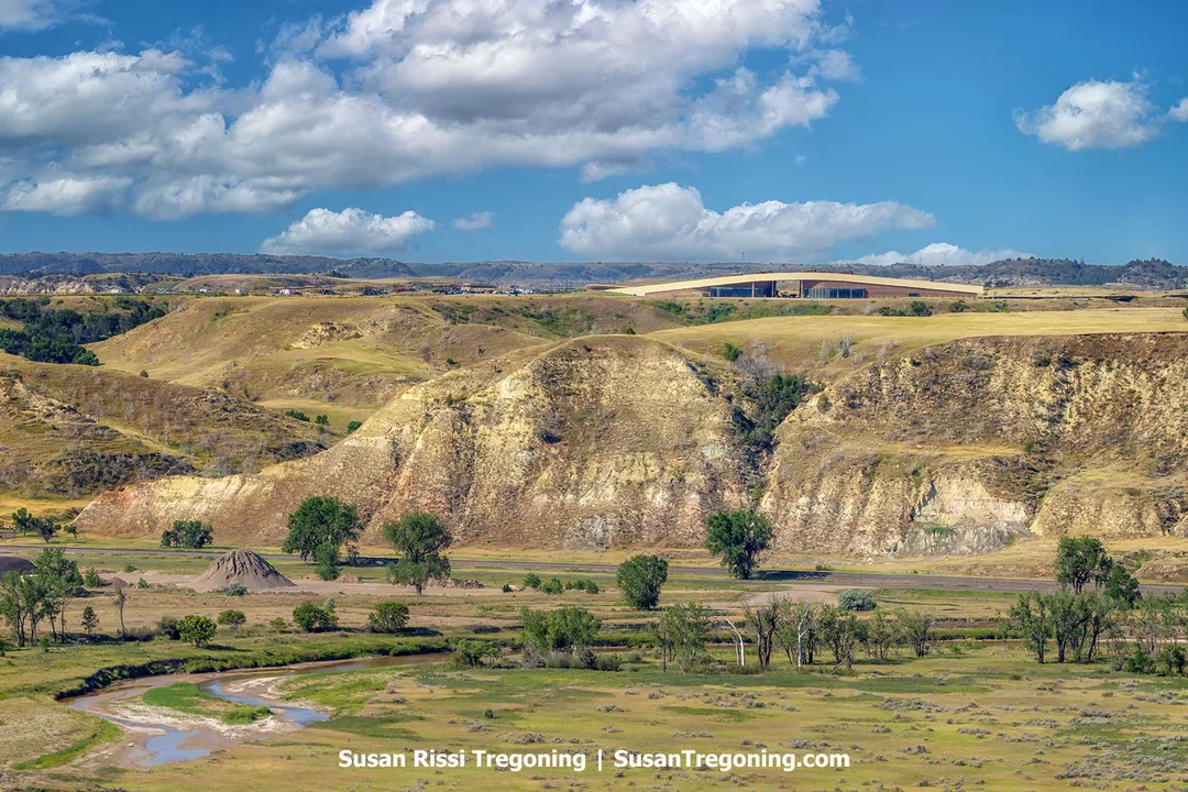 The Theodore Roosevelt Presidential Library in Medora, North Dakota, blends seamlessly into the golden Badlands hills. Its curved roof mirrors the natural contours of the landscape, surrounded by rugged terrain, green valleys, and scattered cottonwoods under a bright blue sky.