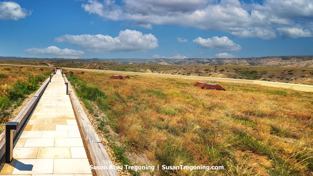 A pale stone walkway stretches through rooftop prairie grasses atop the Theodore Roosevelt Presidential Library in Medora, North Dakota. Small rust‑colored skylights dot the landscape, and the distant Badlands unfold under a bright blue sky, blending architecture seamlessly with nature.