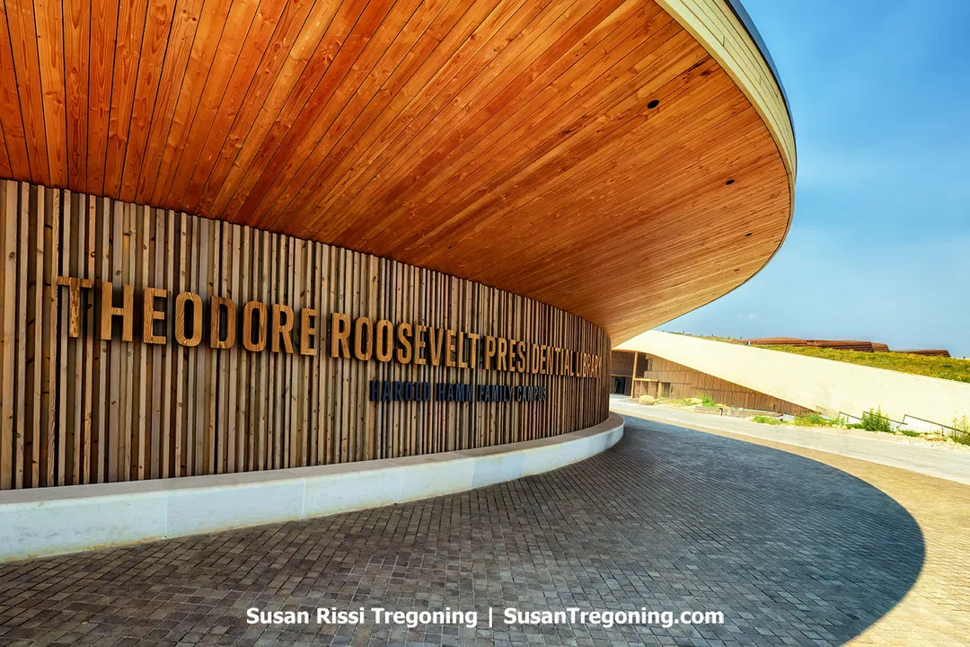 The entrance to the Theodore Roosevelt Presidential Library in Medora, North Dakota, features a sweeping curved wooden facade that blends modern design with natural warmth. Vertical timber slats and bold lettering catch the sunlight, echoing the textures and tones of the surrounding Badlands.
