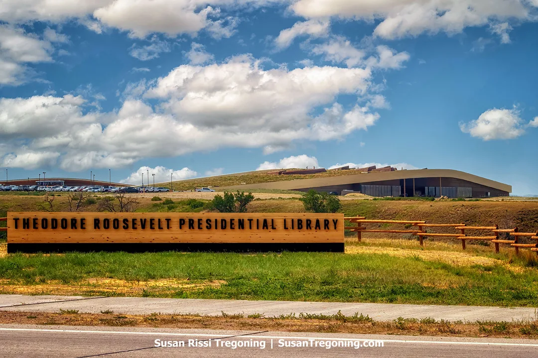 The Theodore Roosevelt Presidential Library sits atop the rolling Badlands near Medora, North Dakota. Its curved, earth‑toned roofline mirrors the surrounding hills, blending modern architecture with the natural landscape under a bright blue sky filled with scattered clouds.